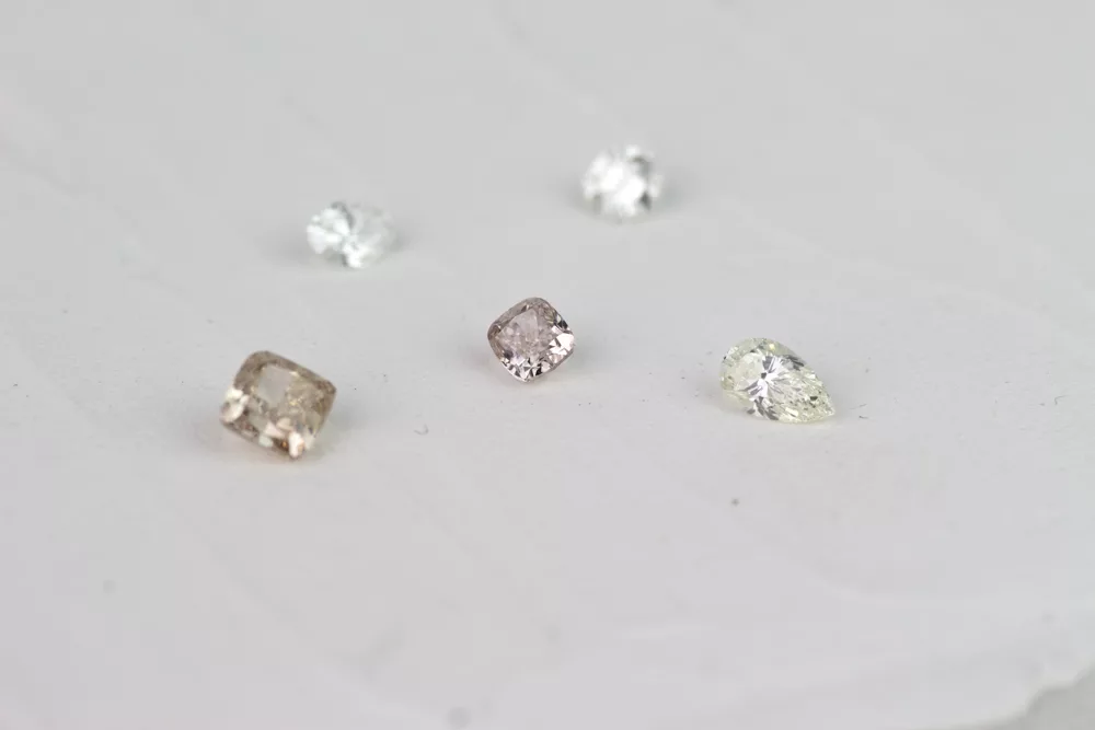 Das Bild zeigt vielfarbigen Diamanten in verschiedenen Schliffen und Farben. Zu sehen sind weiße Diamanten im bekannten Brillantschliff und als Oval, sowie ein brauner und ein champagner Farbener Diamant im Cushionschliff. (Kissenschliff).